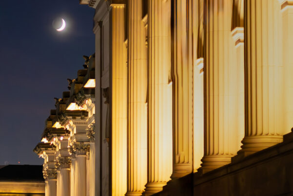 Crescent moon over illuminated classical columns at night, evoking quiet, boundary, and intentional withdrawal.