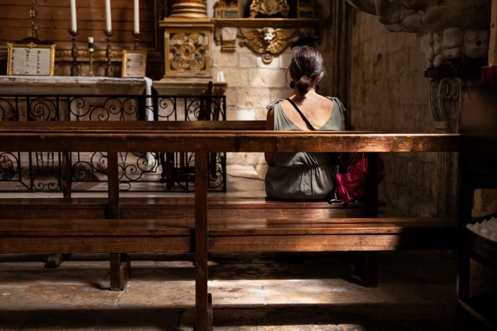 Woman seated alone in a historic church interior with wooden pews, stone walls and warm light.