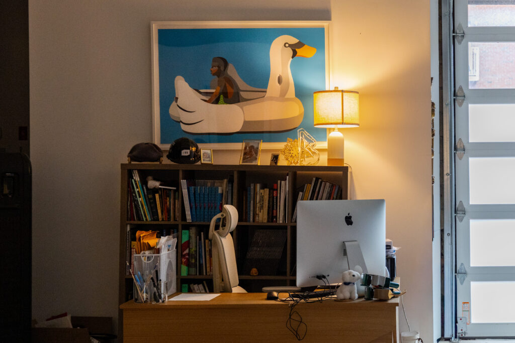 Office corner in Derrick Adams’ Crown Heights studio with books, desk, lamp, computer, and a framed swan artwork.