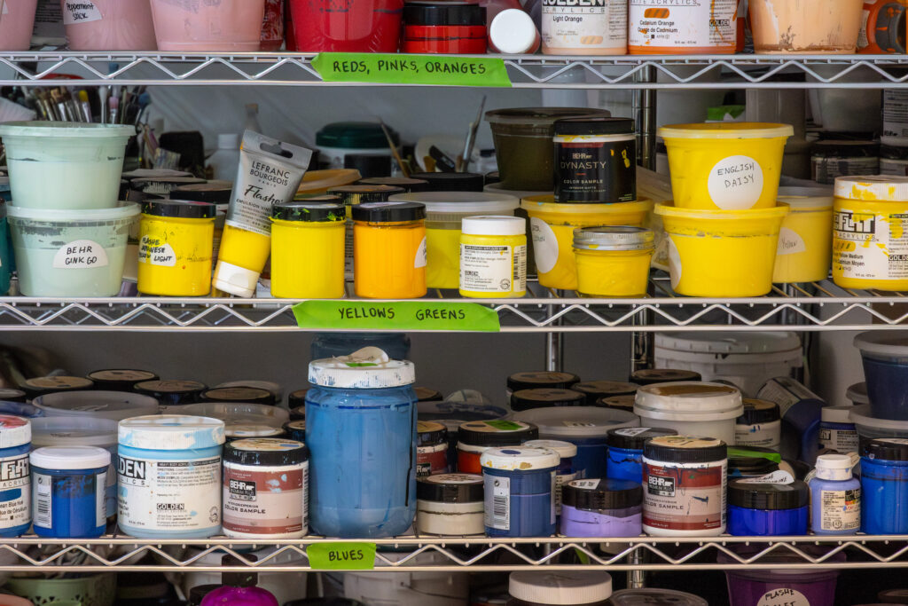 Color-coded shelves of paint in Derrick Adams’ Crown Heights studio labeled reds, pinks, oranges, yellows, greens, and blues.