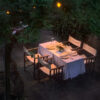 Luxury outdoor dining room at twilight with a table, chairs, garden greenery, and glowing lights.