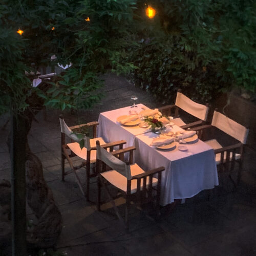 Luxury outdoor dining room at twilight with a table, chairs, garden greenery, and glowing lights.