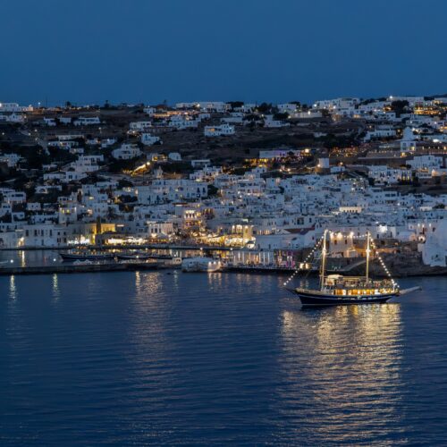 Blue hour view of Mykonos harbor with illuminated boat and whitewashed hillside buildings in Greece