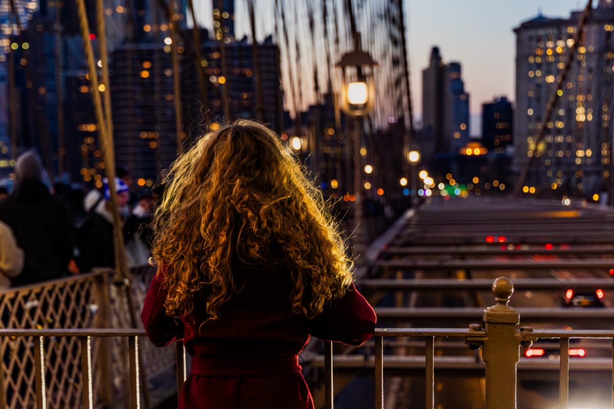 Woman on a New York City bridge at dusk, looking toward city lights, for a guide to where to take visiting foodies in NYC.