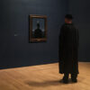 Museum visitor dressed like a bowler man viewing a Magritte painting of a bowler-hatted figure under a crescent moon at SFMOMA.