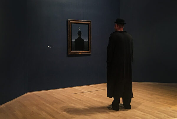 Museum visitor dressed like a bowler man viewing a Magritte painting of a bowler-hatted figure under a crescent moon at SFMOMA.