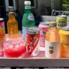 Colorful summer sparkling drinks with bottles, cans and a pink drink over ice on an outdoor tray.