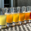 A tasting flight of six iced teas arranged from pale green to deep amber on an outdoor metal table for Dandelion Chandelier’s guide to the best iced teas to buy online.