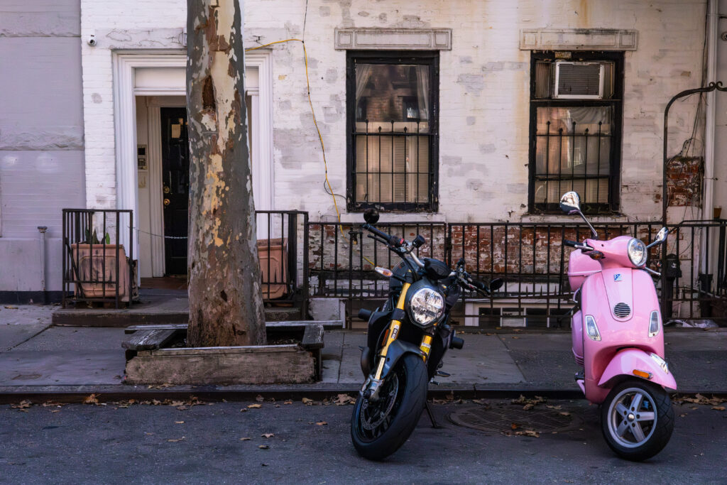 Black motorcycle parked beside a bright pink scooter on a city street, photographed by Pamela Thomas-Graham for romantic gifts with bite.