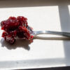 Dark red whole-fruit preserve on a silver spoon in strong morning window light, photographed by Pamela Thomas-Graham.