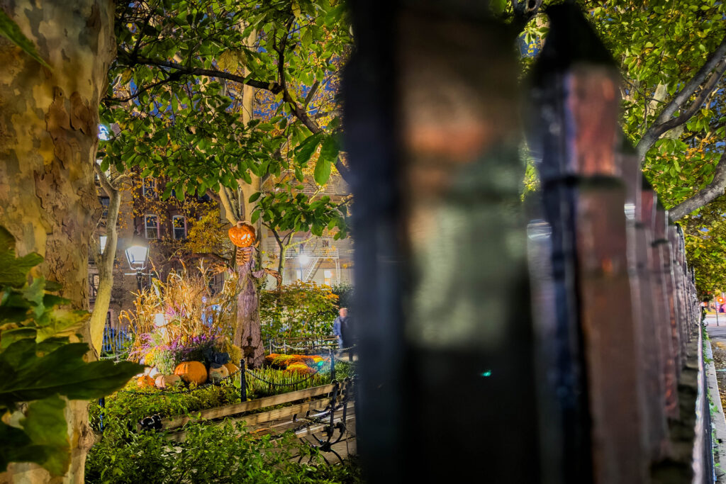 Pumpkin-headed Halloween figure seen through an iron fence at night, photographed by Pamela Thomas-Graham.