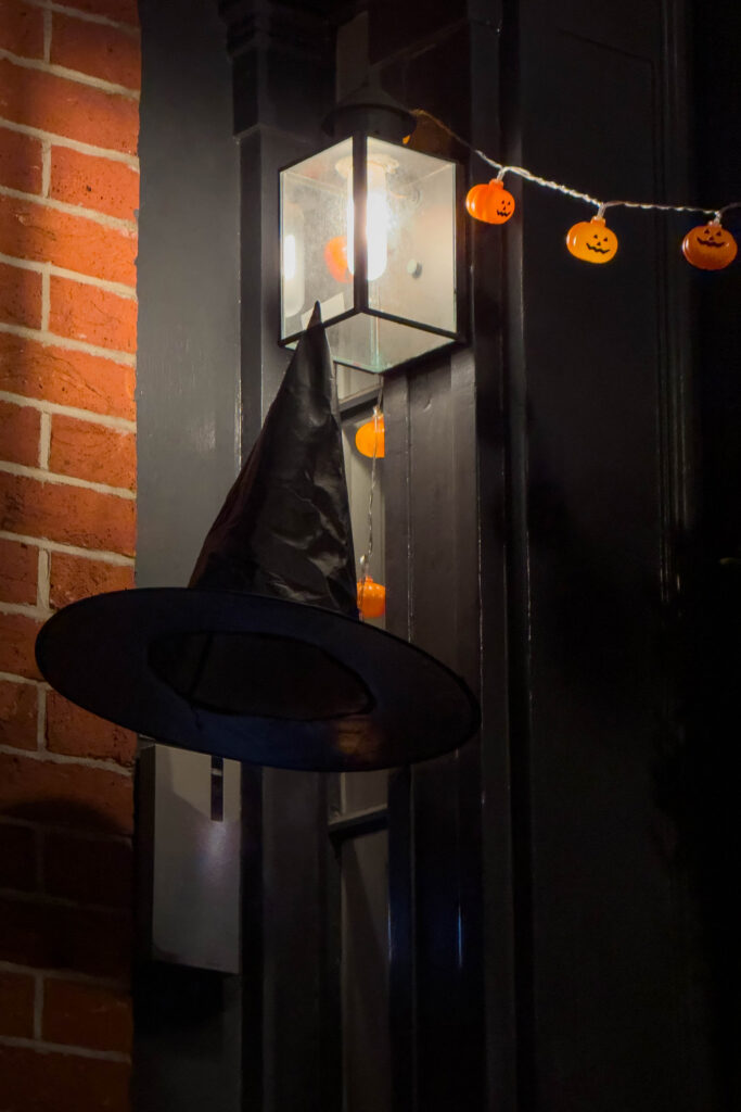 Black witch hat hanging beneath a porch light with small pumpkin lights during Halloween season, photographed by Pamela Thomas-Graham.