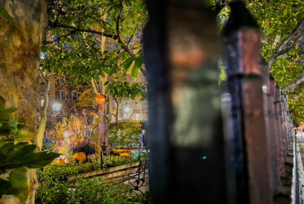 Pumpkin-headed Halloween figure seen through an iron fence at night in New York City, photographed by Pamela Thomas-Graham.