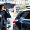 Woman carrying bags under an umbrella on a rainy city street, illustrating power dressing for work with authority, mobility and stamina.