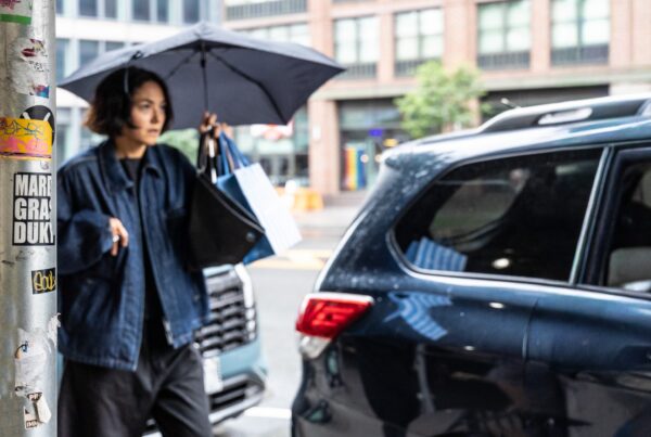Woman carrying bags under an umbrella on a rainy city street, illustrating power dressing for work with authority, mobility and stamina.