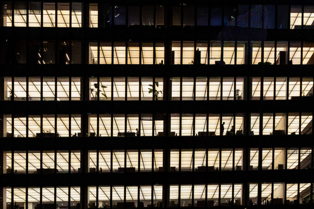 Rows of illuminated office windows at night illustrating the workplace as a system of roles, relationships, and professional expectations.